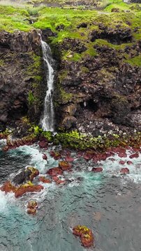 Coastal waterfall flowing from green cliff directly into ocean, Azores islands aerial view with Atlantic waves below