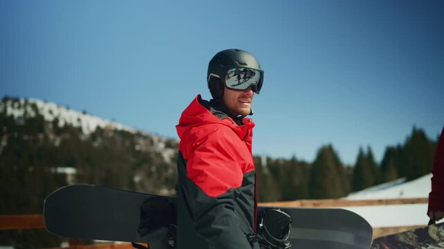 A happy male snowboarder in full gear holds his snowboard on a sunny mountain. The man is enjoying the beautiful winter weather and snowy landscape.
