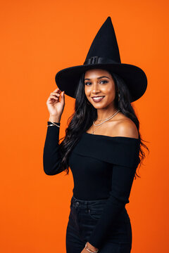Smiling Woman in Black Witch Costume Against Vibrant Orange Background for Halloween