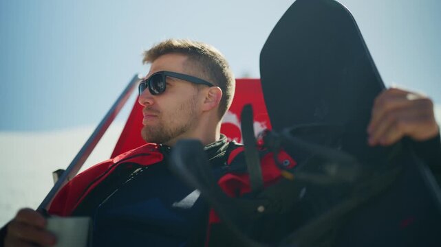 A man in sunglasses and a red jacket relaxes outdoors, holding his snowboard and enjoying the sunshine. He takes a break from snowboarding, looking content in the bright winter landscape.