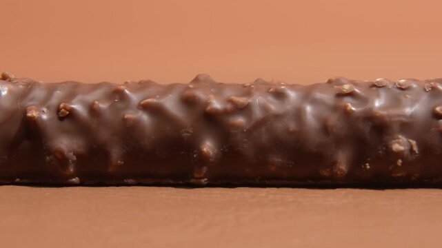 Macro lateral tracking shot moving left along a chocolate coated snack bar with crunchy nut pieces, revealing glossy surface detail against a warm brown studio background.
