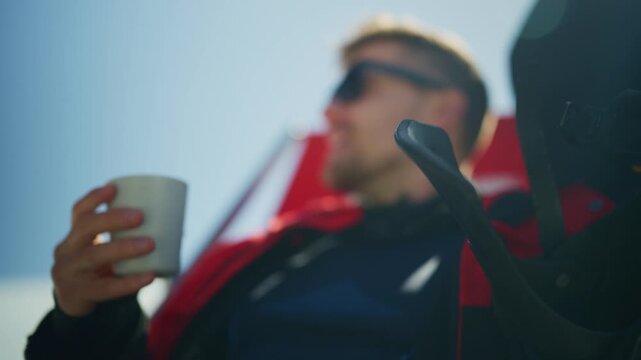 A young man wearing sunglasses relaxes outdoors, enjoying a hot drink on a bright, sunny day. He takes a peaceful break, looking out at the clear blue sky with a content expression.