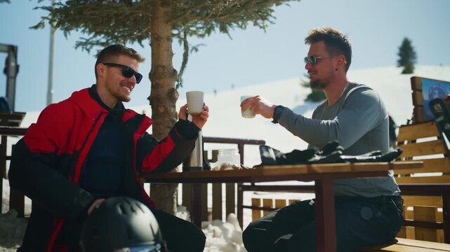 Two male friends are enjoying warm drinks at a mountain ski resort on a sunny winter day. They are clinking their mugs and smiling, taking a relaxing break from skiing activities.