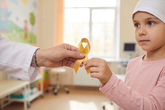  Childhood cancer awareness. Doctor giving a gold ribbon to a little girl patient wearing a medical cap in the hospital room