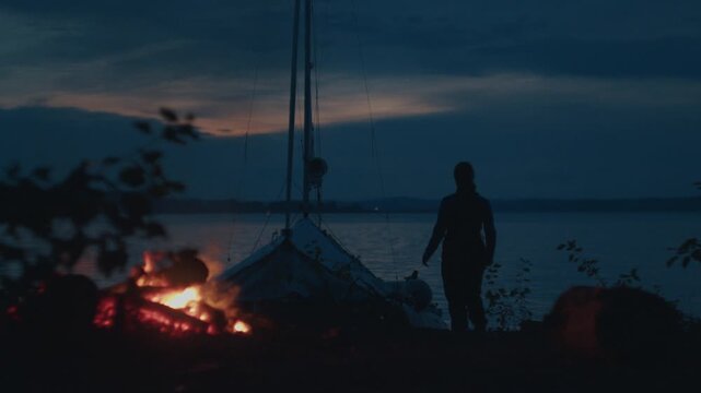 Sailing and camping in the wild. Woman sailor walks on the wild area with sailing boat on the background and bonfire on the foreground