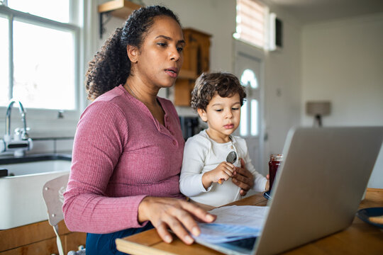 Mother working on laptop with toddler at kitchen table at home