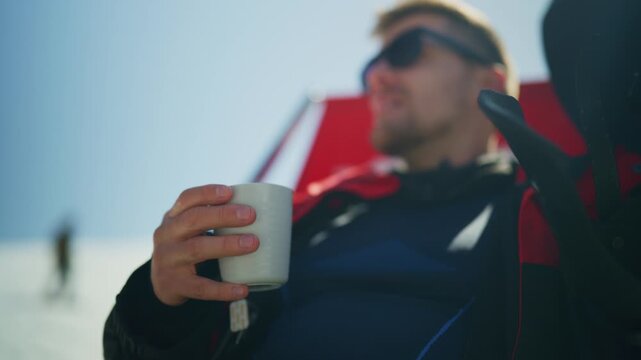 A man in sunglasses relaxes with a warm drink while enjoying the sunny winter outdoors. He takes a peaceful break on a cold day, sipping from a mug.