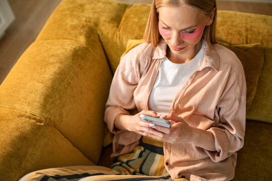 Young woman with eye patches using smartphone on sofa at home