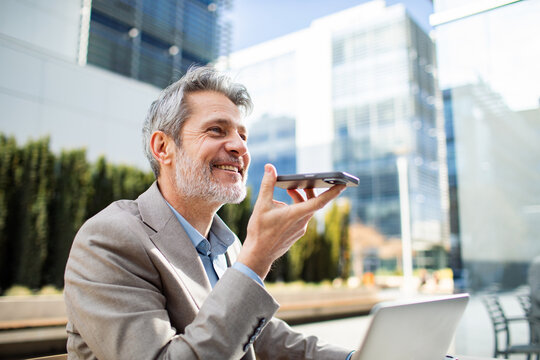Smiling senior businessman using voice assistant on smartphone in modern city office plaza