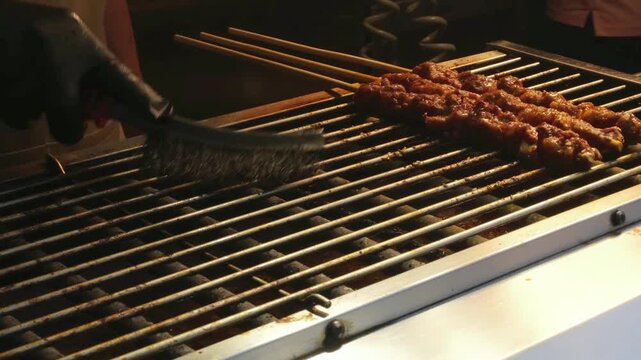worker cleaning grill with brush while cooking meat skewers on hot grill. street food preparation requires clean grill for safe and tasty food in busy restaurant kitchen work zone space
