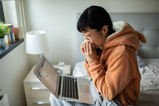 Woman blowing her nose while working on laptop in bed at home