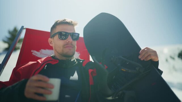 A relaxed male snowboarder enjoys a hot drink while sitting in a deck chair on a sunny mountain. He takes a break from snowboarding, soaking in the winter sun.