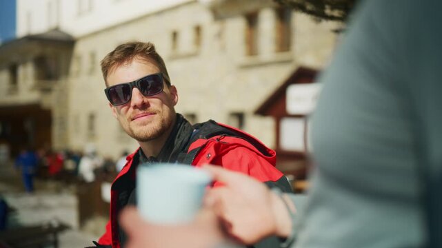 A man in sunglasses shares a warm drink with a friend during winter. They are enjoying hot beverages in a sunny outdoor setting.