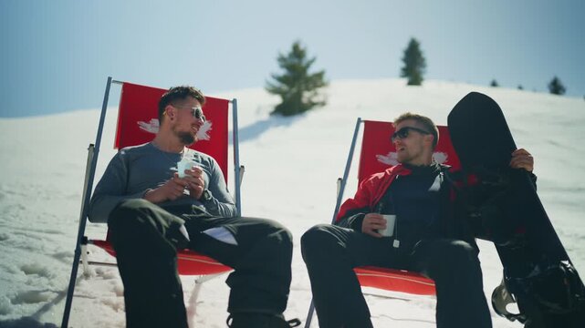Two male snowboarders enjoy a sunny break, relaxing in red deck chairs on a snowy mountain. Friends chat and laugh, savoring their time together after winter sports.