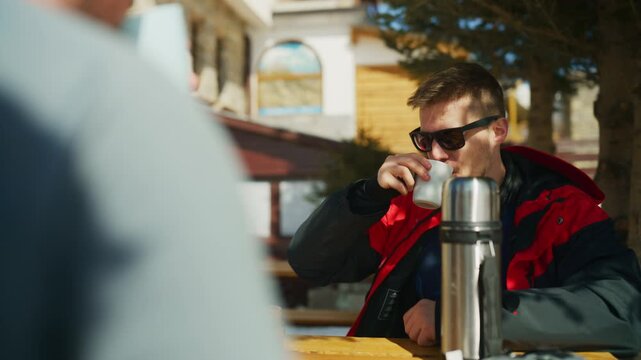 A man in sunglasses sips a warm drink and laughs with a friend at an outdoor cafe. Two happy friends enjoy a casual conversation and hot beverages on a sunny winter day.