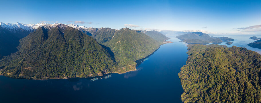 A deep blue fjord winds between forested mountains and snowy peaks, calm waters stretching toward the open sea.