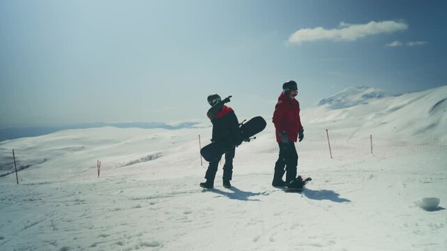 Two snowboarders stand on a snowy mountain, preparing to descend the slope. They are enjoying the winter sports and the beautiful landscape on a bright, sunny day.