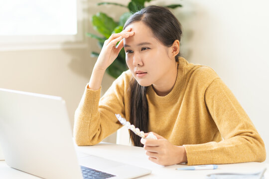 Sick asian young woman holding a blister pack of pills and touching her forehead during a telehealth consultation on a laptop, explaining her headache symptoms to an online doctor.