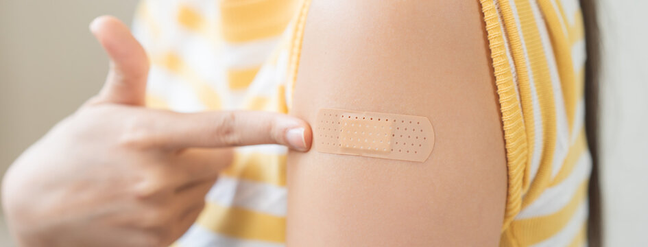 Close up of a young woman pointing at an adhesive bandage on her arm after receiving a vaccine injection, representing healthcare, virus immunity protection, and prevention.