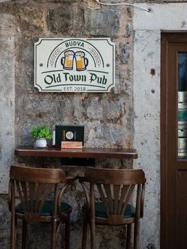 Two wooden chairs are set at a small outdoor counter beneath the sign of the Old Town Pub in Budva, Montenegro on August 9, 2026