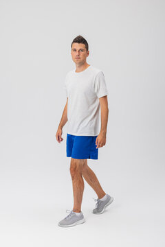 Full-body portrait of a trim athletic man walking to the side against an isolated white studio background, wearing a light grey t-shirt and blue athletic shorts.