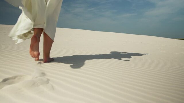 Woman's bare feet walking on sand dunes as her white dress flutters in the wind leaving footprints in the white desert sand