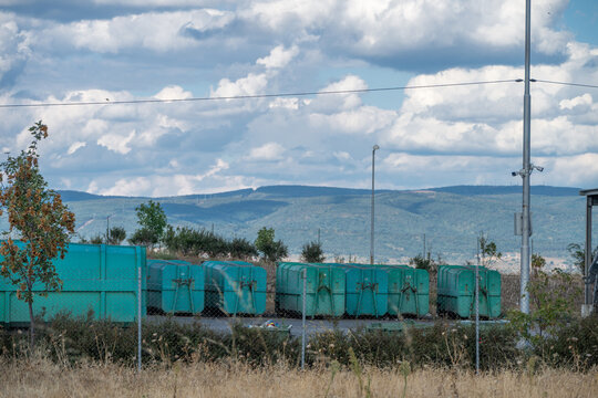 A row of large green industrial waste compactors and roll-off containers sits parked at a municipal recycling depot against a backdrop of distant hills and a cloud-filled sky.