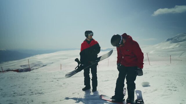 Two snowboarders are preparing their equipment on a sunny, snow-covered mountain. They are getting ready for an exhilarating day of winter sports and adventure.