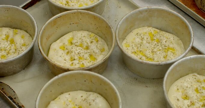 hands placing cherry tomato slices onto risen raw dough rounds that are dotted with olive oil and sprinkled herbs inside round metal molds on worn baking tray