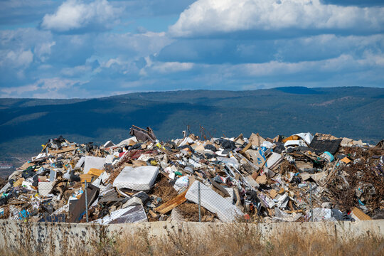 A wide mound of bulky domestic waste, discarded mattresses, and demolition debris fills an open landfill behind concrete barrier walls, set against distant blue hills and a cloud-swept sky.