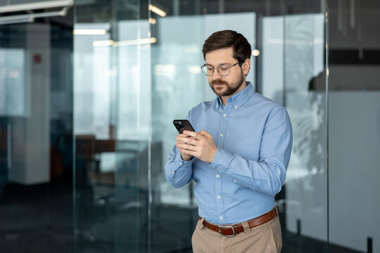 A young, serious businessman stands in an office and uses a mobile phone