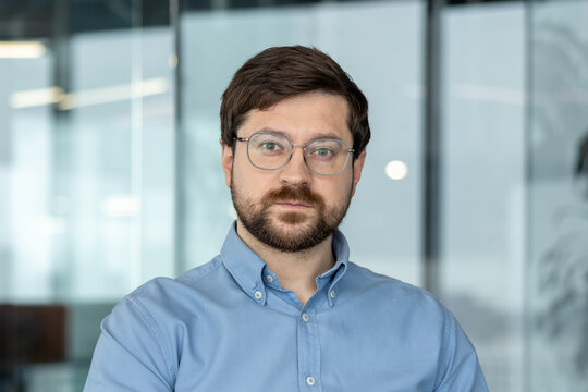 A close-up portrait of a young man in glasses and a shirt, located in an office setting, looking seriously at the camera