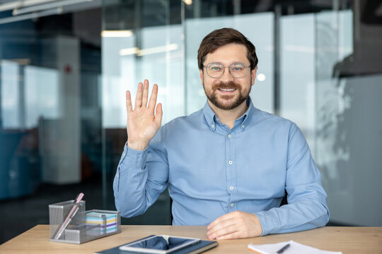 A portrait of a smiling young man in an office; he is sitting at his desk, waving hello, and starting a remote conversation on camera