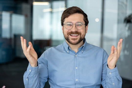 A portrait of a smiling young man in an office setting, speaking to the camera, presenting, and gesturing with his hands