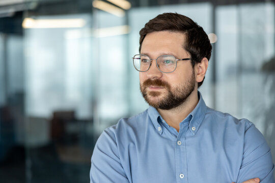 A close-up photo of a young man wearing glasses and a shirt in an office setting, smiling and looking off to the side