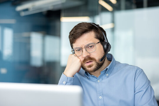 A dejected and tired young man sits at an office desk in front of a laptop, resting his head on his hand and looking at the screen