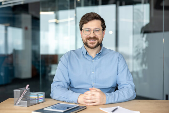 A portrait of a young, smiling man wearing glasses and a shirt; he is sitting at an office desk with his hands clasped, looking confidently at the camera