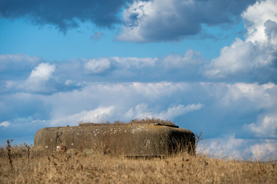 An weathered concrete military pillbox bunker rests on a dry, grassy hill ridge beneath a dramatic sky filled with thick white and dark clouds.