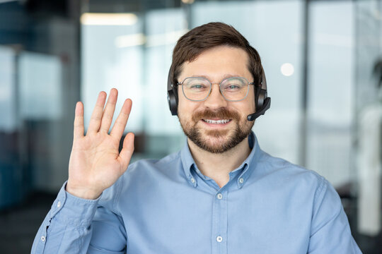 A smiling young man wearing a headset conducts an online video meeting while sitting at a desk in an office, greeting the camera