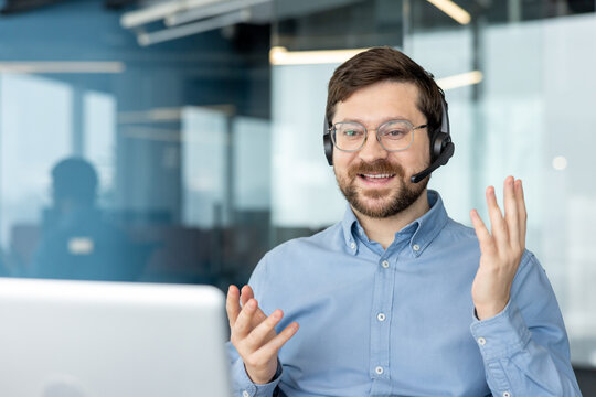 A smiling young man wearing a headset is conducting a video meeting on a laptop; he is sitting at a desk in an office, talking and gesturing with his hands