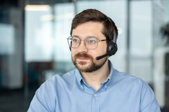 A close-up photo of a serious young man wearing a headset, glasses, and a blue shirt, sitting in an office and looking intently off to the side