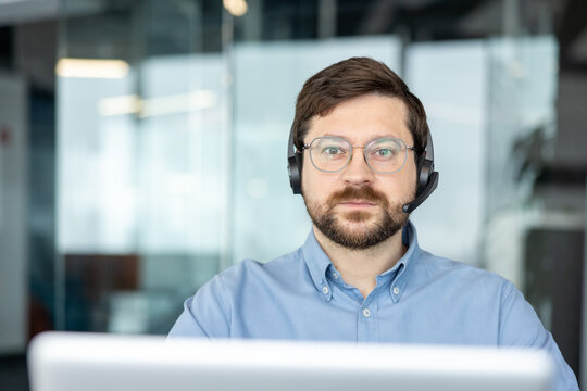 A close-up portrait of a young man wearing a headset, sitting at a desk with a laptop in an office and looking seriously at the camera