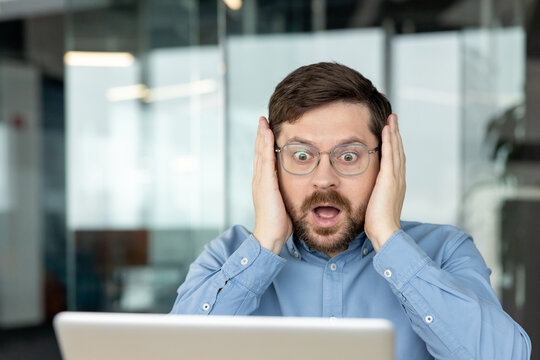 A frightened young man sits in an office, holding his face with his hands and looking anxiously at his laptop screen as he reads the news, receives a message, or watches a video