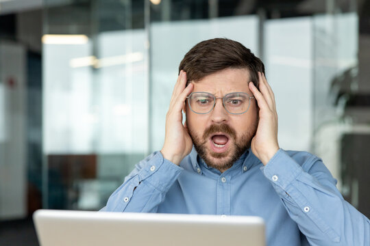 A close-up photo of a young, frustrated businessman sitting at an office desk, holding his head, and looking at a laptop screen