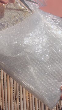 woman pulls clear bubble wrap tight around picture corner while standing outdoors next to bamboo fence, showing careful protective packing hands gripping wrap