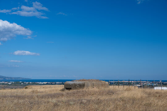 An old concrete military bunker pillbox overlooks a dry grassy field, distant coastal towns, and the deep blue sea under an expansive clear sky.