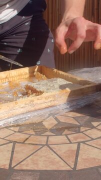 close-up view of hands lowering rectangular honeycomb piece onto white plate, thick amber honey strands stretching and dripping under sunlight on patio at noon