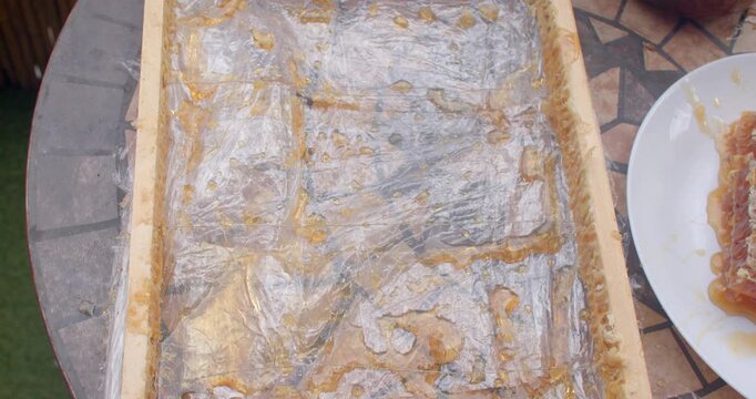 top-down oblique view of wooden honey board covered in clear plastic wrap with smeared amber honey pools after extraction, spoon removed between shots soon pool
