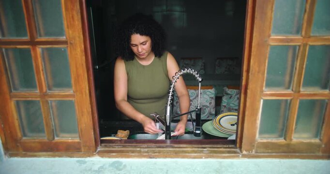 Woman washing dishes at kitchen sink, seen through wooden window frame, serene moment in a cozy home environment, focused on daily routine