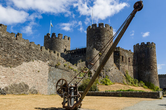 Ancient stone fortification walls and round towers under blue sky with clouds, historic iron winch mechanism on grass in foreground.